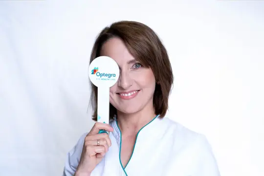 Uśmiechnięta kobieta w białym fartuchu zasłania jedno oko tabliczką z logo "Optegra Eye Health Care". To symbol nagłej poprawy wzroku.