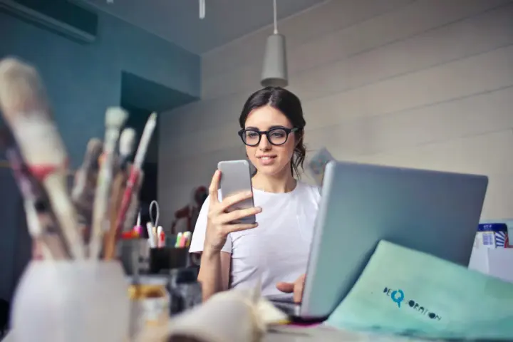 Kobieta w okularach do komputera, zastanawia się czy warto je nosić, przeglądając coś na telefonie.