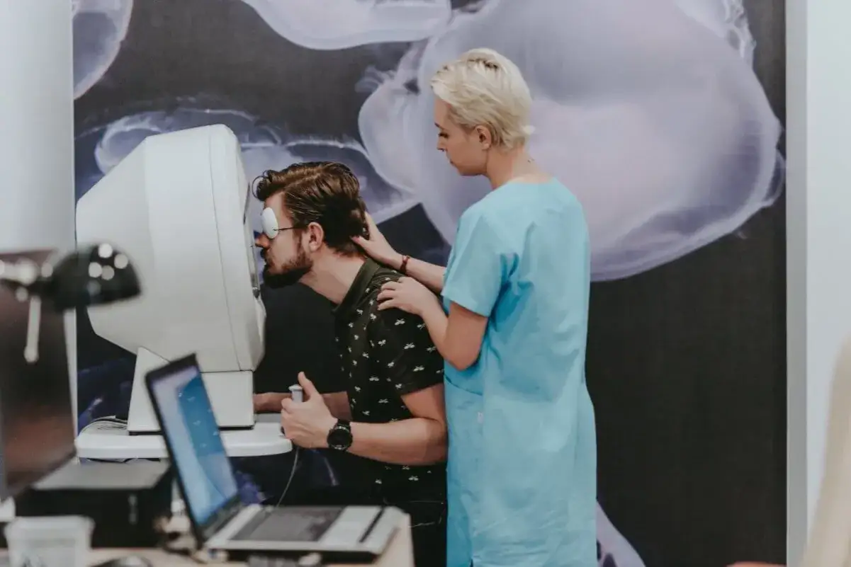 Mężczyzna z zasłoniętym okiem bada swoje widzenie tunelowe na specjalistycznym urządzeniu, podczas gdy optometrystka go wspiera.